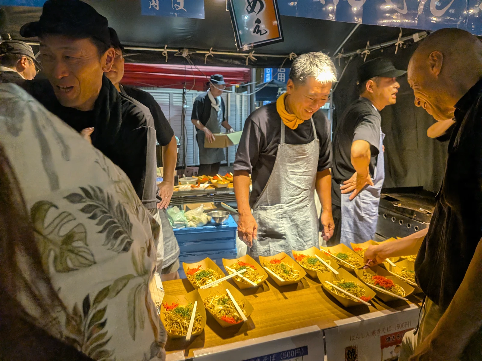 左側と右側に笑顔の客、中央に笑顔の作り手、舟皿の焼きそばが並ぶ露店