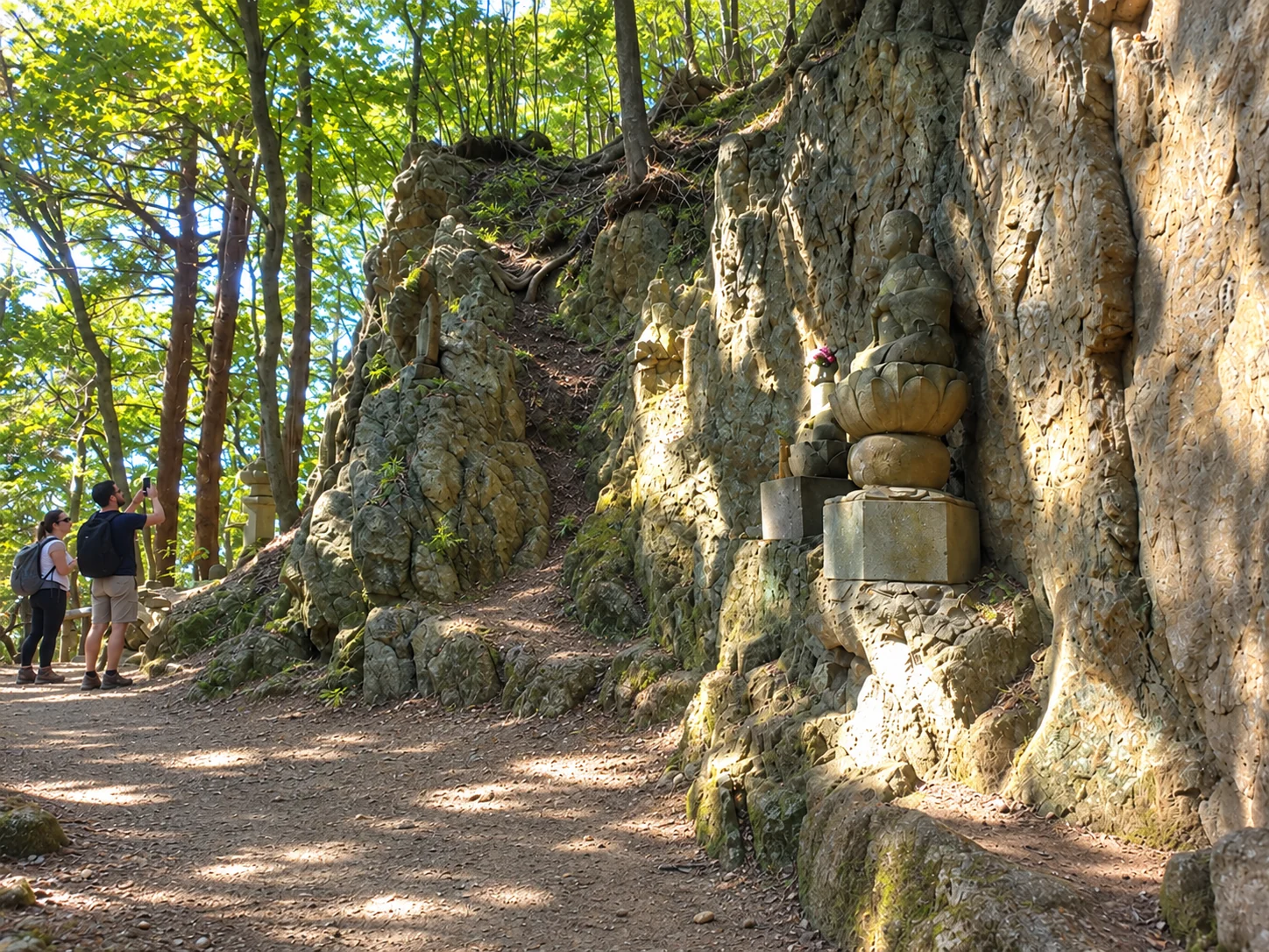 Visitors walking beside a sunlit rock wall lined with stone figures